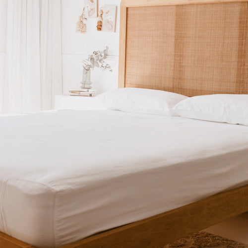 queen bed with rattan had board and white curtains with white pillowcases and white fitted sheet, showing the corner of the fitted sheet has an additional stretch panel
